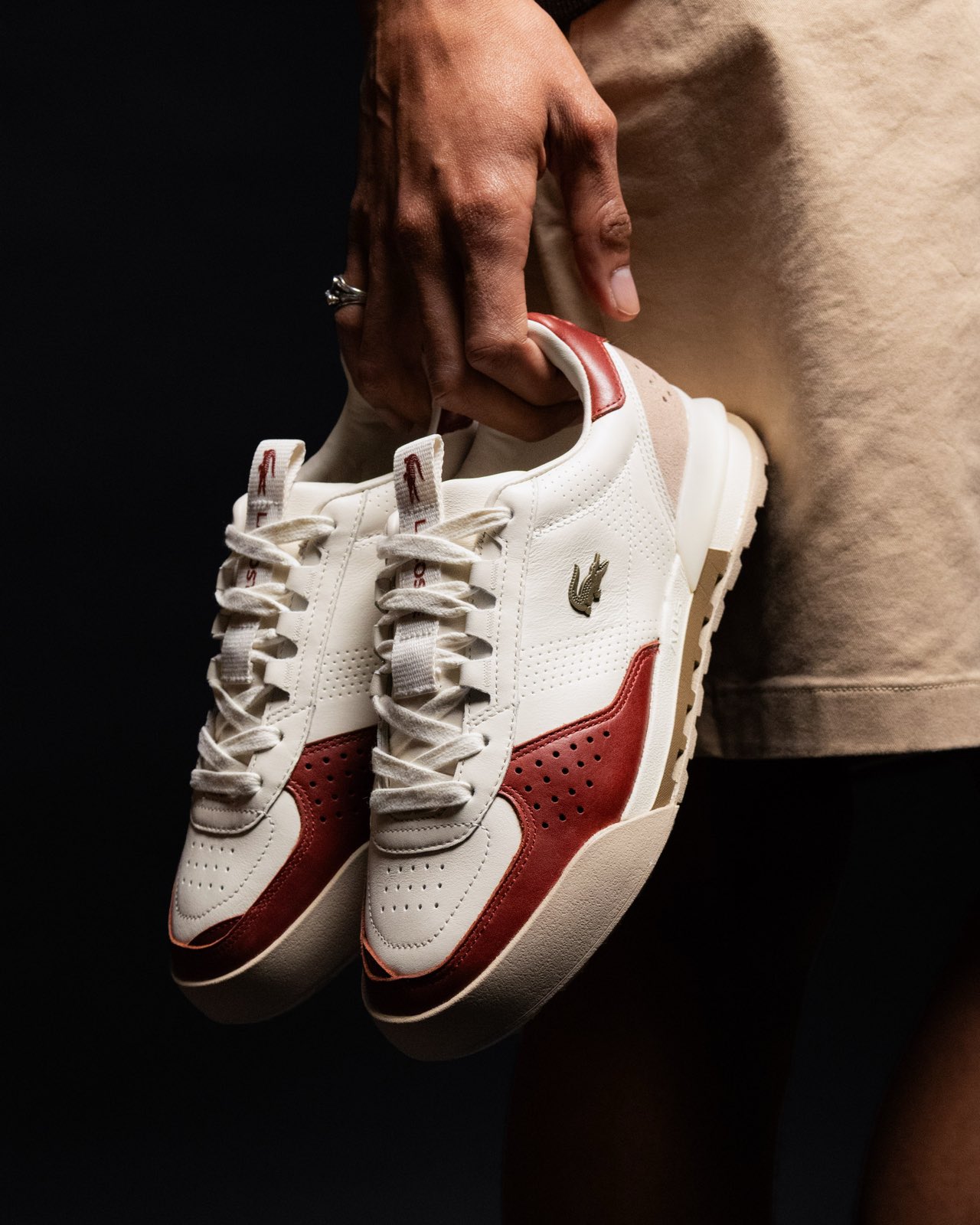 Hands holding a red and cream Lacoste court sneaker, black studio